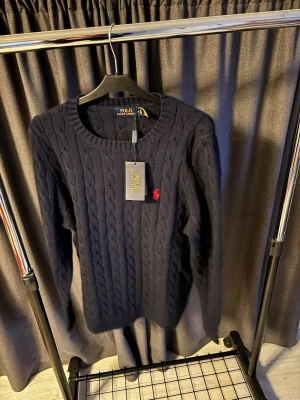 Mörkblå kabelstickad tröja Polo Ralph Lauren - Kabelstickad mörkblå tröja från Polo Ralph Lauren med röd broderad log typ. Perfekt för dig som gillar en stillren look| Ny med prislapp| storlek M| Hör gärna av er vid funderingar 