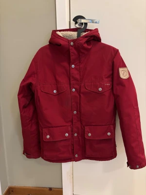 Röd parkajacka från Fjällräven - Röd parkajacka från Fjällräven med huva och fyra framfickor med tryckknappar. Jackan har ett klassiskt Fjällräven-märke på ärmen och mysigt foder i luvan. Perfekt för kyliga dagar och outdoor-äventyr.