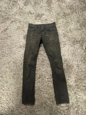 Svarta skinny jeans D02 M/SK-LW YSL - Säljer ett par svarta skinny jeans med låg midja och klassisk femficksdesign. Jeansen har en diskret slitning vid ena fickan och är perfekta för en avslappnad streetstil. Passar dig som gillar smal passform och mörka tvättar.