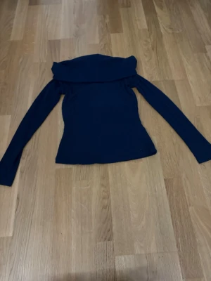 Marinblå offshoulder topp Gina Tricot - Trendig marinblå offshoulder topp med långa ärmar och figurnära passform. Toppen har en bred krage som vilar snyggt över axlarna och är tillverkad i ett mjukt, stretchigt material som ger en bekväm känsla.