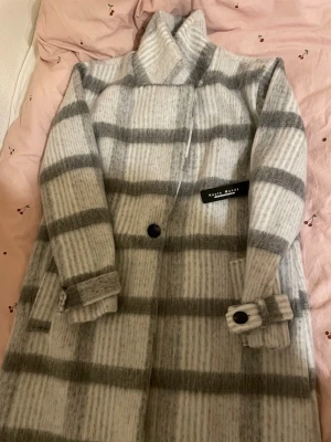 Gråvit rutig kappa från House of Dagmar - Snygg lång kappa i grått och vitt från House of Dagmar. Kappan har breda horisontella ränder, stora svarta knappar och hög krage. Tillverkad i ett mjukt, ulligt material som känns lyxigt och varmt. Perfekt för att lyfta din outfit under kyliga dagar.