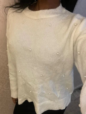 Vit stickad tröja med pärlor - Säljer en vit stickad tröja med dekorativa pärlor på fram- och ärmsidorna. Tröjan har rund halsringning, långa ärmar och ribbade muddar. Perfekt för dig som gillar en enkel men ändå trendig look.
