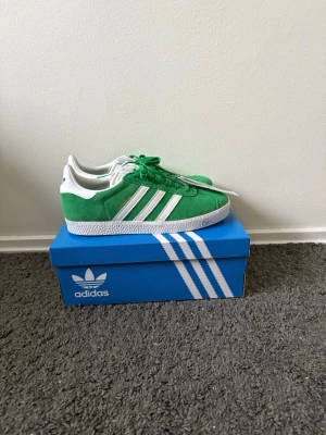 Gröna Adidas Gazelle sneakers - Säljer ett par gröna Adidas Gazelle sneakers med klassiska vita ränder på sidan och vit sula. Ovandelen är i mocka och skorna har gröna skosnören samt vit hälflik med Adidas-logga. Perfekta för dig som vill sticka ut med en retrovibe.