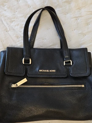 Svart handväska Michael Kors läder - Snygg svart handväska från Michael Kors i strukturerat läder med guldfärgade detaljer. Väskan har dubbla handtag, en framficka med dragkedja och logga i guld på framsidan. Klassisk och stilren design som funkar till många looks.