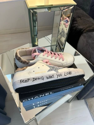 Golden Goose sneakers med rosa snören - Golden Goose sneakers i vitt läder med rosa snören, silverglittrig stjärna på sidan och leopardmönstrad häl. Texten 'KEEP DOING WHAT YOU LOVE' på sulan ger en personlig touch. Perfekta för dig som vill sticka ut med coola detaljer.