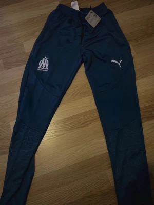 Marinblå träningsbyxor Puma OM - Snygga marinblå träningsbyxor från Puma med Olympique de Marseille-logga och vit Puma-logga på benen. Byxorna har smal passform, dragsko i midjan och diskret mönster på nedre delen av benen. Perfekta för träning eller chill.