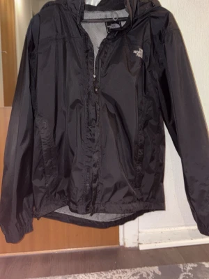 Svart regnjacka från The North Face - Snygg svart regnjacka från The North Face med huva och dragkedja framtill. Jackan har meshfoder på insidan för extra ventilation och elastiska muddar vid ärmsluten. Perfekt för regniga dagar och enkel att packa ner i väskan.