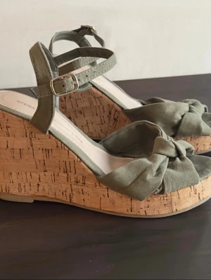 Sfera 36 Olivgröna kilklackar sandaler med rosett - Färg: militärgrön. Strl: 36. Klackhöjd baktill: 9cm & 2,5cm fram. Snygga olivgröna sandaler från Sfera med kilklack i kork och dekorativ rosett framtill. Sandalerna har dubbla remmar med spänne runt ankeln och öppen tå. Ovandelen är i mjukt tyg som ger en trendig och bekväm känsla.