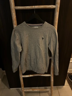 Grå sweatshirt från U.S. Polo Assn. - Klassisk grå sweatshirt från U.S. Polo Assn. med rund halsringning och diskret broderad logga på bröstet. Tröjan har ribbade muddar vid ärmslut och nederkant, perfekt för en avslappnad och stilren look.