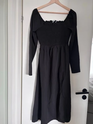 Svart långärmad klänning från NA-KD - Svart långklänning från NA-KD med smockad överdel och långa ärmar. Klänningen har en söt volangkant vid urringningen och en figurnära passform upptill som faller ut i en vidare kjol. Perfekt för dig som gillar stilrena och trendiga plagg.