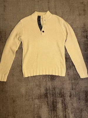 Beige stickad tröja med knappar - Säljer en beige stickad tröja från NN07 med ribbad hög krage och  fem bruna knappar framtill. Tröjan har lång ärm, ribbade muddar och ett snyggt strukturmönster. Perfekt för dig som gillar stilrena och bekväma plagg.