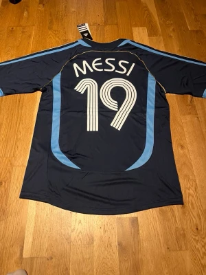 Argentina 2006 long sleeve Messi #19 - Mörkblå långärmad fotbollströja från Adidas med ljusblå detaljer, nummer 19 och Messi på ryggen. Tröjan har Argentinas landslagsemblem på bröstet och vita Adidas-loggor. Perfekt för dig som älskar fotboll och vill stötta Argentina.