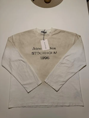 Vit långärmad topp Acne Studios - Vit långärmad topp från Acne Studios med tryck 'STOCKHOLM 1996' framtill. Klassisk rak passform och rund halsringning. Tillverkad i mjuk bomull, perfekt för en clean och stilren look.