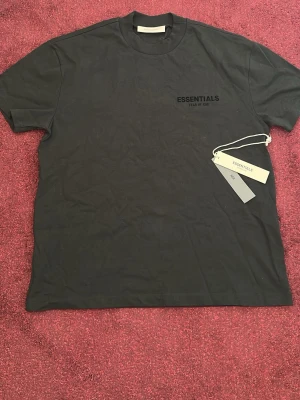 Svart Essentials Fear of God t-shirt - Snygg svart t-shirt från Essentials Fear of God med tryck på både bröstet och ryggen. Klassisk rund hals och korta ärmar. Tillverkad i mjuk bomull för skön känsla och clean look. Perfekt för dig som gillar streetwear och vill ha en enkel men stilren tee.