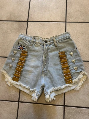 Ljusblå denimshorts med nitar och detaljer storlek S/M - Unika ljusblå denimshorts från Famous med hög midja, råa fransade kanter och coola nitar samt metallspännen på sidorna. Perfekta för dig som vill sticka ut i sommar! Midja 68 cm.