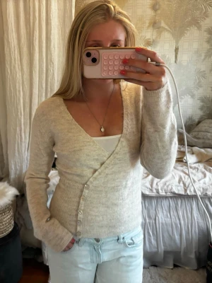 Beige omlottkofta från Gina Tricot - Snygg beige omlottkofta från Gina med v-ringning och långa ärmar. Koftan har ribbad midja och knäppning med pärlemorknappar längs sidan. Perfekt att styla med jeans eller kjol för en chill look.