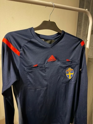 Blå långärmad domartröja Adidas - Mörkblå långärmad domartröja från Adidas med röda detaljer och två bröstfickor. Svenska fotbollsförbundets märke på bröstet och Fair Play SvFF-tryck på ärmen. Perfekt för dig som dömer fotbollsmatcher och vill ha officiell stil.