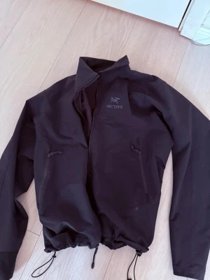 Svart vindjacka från Arc'teryx - Snygg svart vindjacka från Arc'teryx med dragkedja framtill, två snedställda fickor med dragkedja och justerbar dragsko i nederkant. Jackan har hög krage och diskret logga på bröstet. Perfekt för blåsiga dagar och outdoor-äventyr.