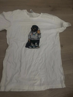 Vit t-shirt med tryck av figur - Vit t-shirt med korta ärmar och ett stort tryck framtill av en tecknad figur med mask och hoodie. T-shirten har en avslappnad passform 
