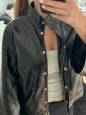 Svart glansig skinnjacka (fejk)  - Snygg svart skinnjacka med glansig finish och hög krage. Jackan har dragkedja och tryckknappar framtill för en cool look. Rak passform och långa ärmar. Perfekt jacka till våren och sommaren då den är ganska tunn. 💞💞💞(Det finns en reva inuti jackan på ryggen, den syns inte när man har på sig  jackan). 