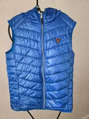 Blå dunväst med huva från Guess - Snygg blå dunväst från Guess med huva och dragkedja framtill. Västen har ett quiltat mönster och Guess-logga på bröstet. Perfekt för lager på lager och ger en sportig vibe till din outfit.