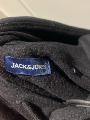 Svart hoodie från Jack & Jones - Svart hoodie från Jack & Jones med vit logga på bröstet. Klassisk känguruficka framtill och justerbar huva med snörning. Perfekt för en avslappnad och stilren look.
