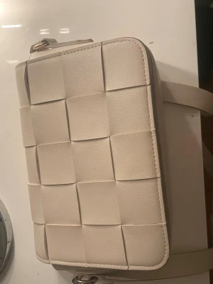 Beige axelväska med flätad design - Snygg beige axelväska med rektangulär form och flätad framsida. Väskan har justerbar axelrem och är tillverkad i konstläder. Perfekt för dig som gillar stilrena accessoarer med modern twist.