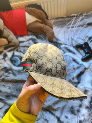 Gucci keps med GG-mönster - Snygg beige keps från Gucci med klassiskt GG-monogram över hela kepsen. Kepsen har en böjd skärm och en grön-röd rand på sidan, typisk för Gucci. Materialet är canvas med justerbart spänne bak. Perfekt för dig som vill sticka ut med en lyxig accessoar.