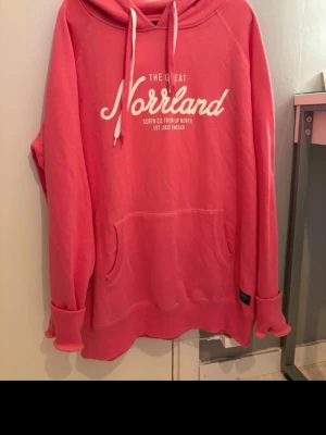 Rosa hoodie från SQRTN Company - Snygg rosa hoodie från SQRTN Company med vit text 'Norrland' på bröstet och känguruficka framtill. Hoodien har dragsko i huvan och är tillverkad i mjukt bomullsmaterial. Perfekt för dig som gillar streetwear och vill ha något bekvämt och stilrent.