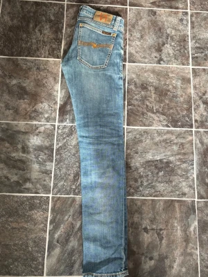 Ljusblå jeans från Nudie - Säljer ett par ljusblå skinny jeans från Nudie Jeans med klassisk femficksdesign och slitningar på låren. Jeansen har en smal passform och är tillverkade i mjuk denim med snygga kontrastsömmar och läderpatch bak i midjan.