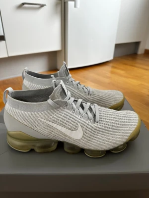 Nike Air Vapormax Flyknit grå - Snygga Nike Air Vapormax Flyknit sneakers i ljusgrått med stickad ovandel och genomskinlig, bubblig sula. Skorna har en vit swoosh på sidan och gråa skosnören. Ovandelen är i ett mjukt och flexibelt material som ger en modern look.