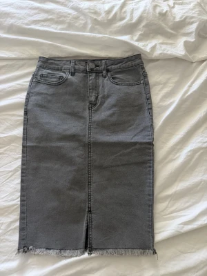 Grå denimkjol med rå kant - Snygg grå pennkjol i denim med klassiska femficksdetaljer och rå, fransad nederkant. Kjolen har slits framtill och bälteshällor i midjan. Perfekt för en trendig och avslappnad look.