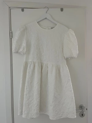 Vit kort klänning med puffärm - Superfin vit kort klänning med puffärmar och rund hals. Klänningen har ett subtilt blommigt mönster i tyget och en lätt struktur. Perfekt för student och sommarens tillfällen och har en söt, feminin siluett.