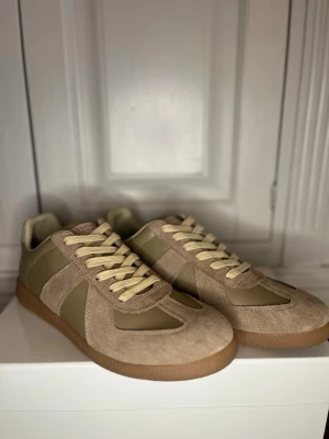 Beige sneakers från Maison Margiela - Snygga sneakers i beige mocka och olivgrönt läder från Maison Margiela. Klassisk low-top modell med krämvita snören och gummisula. Diskret logga på plösen och stilren design som passar till det mesta. Kan sänka pris