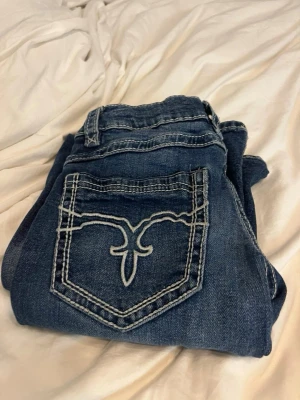 Mörkblå bootcut jeans med brodyr - Snygga mörkblå bootcut jeans med vit brodyr på bakfickorna och markerade sömmar. Jeansen har klassisk femficksdesign och lätt utsvängda ben. Perfekta för dig som gillar en retrostil och vill sticka ut lite extra.