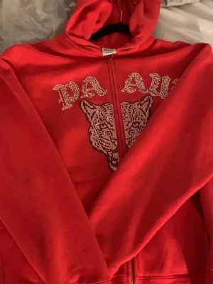 Röd PA AW hoodie med strass - Snygg röd hoodie med dragkedja från PA AW. Framsidan har ett stort kattmotiv och text i strass, samt fickor och justerbar huva med snören. Perfekt för dig som gillar statement-plagg och vill sticka ut lite extra.