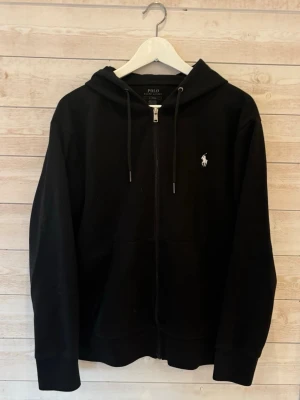 Fet Zip Hoodie  - Svart hoodie med dragkedja från Klassisk design med huva och justerbara snören, samt den ikoniska broderade loggan på bröstet. För en skön och avslappnad stil. 