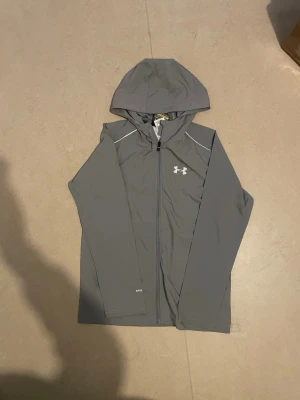 Grå vindjacka från Under Armour - Snygg grå vindjacka från Under Armour med huva och hel dragkedja framtill. Jackan har vita detaljer vid axlarna och logga på bröstet. Tillverkad i lätt och funktionellt material som passar perfekt för blåsiga dagar.