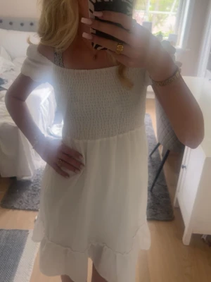 Vit  sommarklänning - Söt vit kortklänning med offshoulder och smockad överdel. Klänningen har volang nertill och är perfekt för varma dagar. Tillverkad i ett lätt och luftigt material som känns skönt mot huden. Fin till skolavslutning eller till sommaren. 