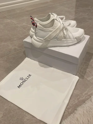 Vita Moncler sneakers med meshdetaljer - Stilrena vita sneakers från Moncler med meshpaneler och detaljer i läder. Skorna har en chunky sula och Monclers signaturrand på hälen. Perfekta för dig som gillar en sportig och modern look.