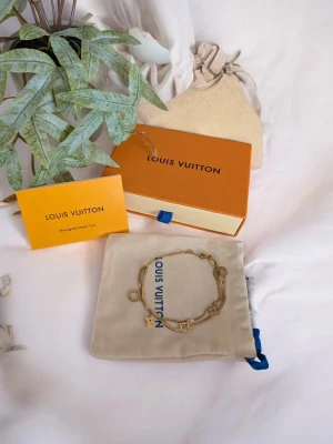 Iouis Vuitton Armband  - Design: Ett sofistikerat armband med dubbla kedjor – en nätt klassisk länk kombinerad med en platt "snake"-kedja. Dekoration: Prydd med flera hängande berlocker i form av geometriska figurer och fyrbladiga blommor. Funktion: Justerbar passform med ett praktiskt klolås. Tillbehör: Säljes som ett komplett set med en orange märkesask, ljus dammpåse och ett "Monogram World Tour"-infokort.