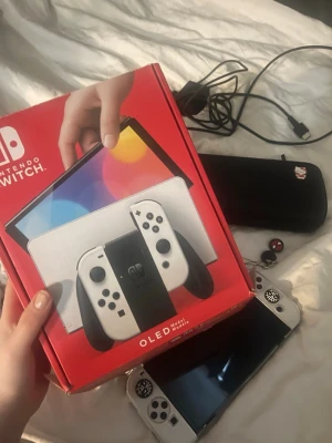 Nintendo Switch OLED vit konsol - Nintendo Switch OLED i vit med matchande Joy-Con-kontroller. Konsolen har en stor OLED-skärm, svart och vit dockningsstation samt tillbehör som laddare, handkontrollshållare och ett svart fodral med dekaler. Perfekt för gaming hemma eller på språng. Köpte den januari 2026. Har knappt spelat :) 