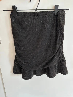Svart glittrig kortkjol med volang - Snygg svart kortkjol med glittriga detaljer och volang nertill. Kjolen har en omlott-look och lätt rynkning på ena sidan. Perfekt för dig som vill sticka ut lite extra. Ingen synlig logga eller märke.