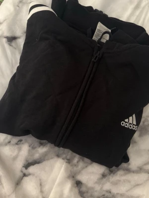 Svart ziptröja från Adidas - Svart ziptröja från Adidas med vit logga på bröstet och vita detaljer på axeln. Tröjan har dragkedja framtill och är tillverkad i mjukt material, perfekt för träning eller chill. Klassisk sportig look med bekväm passform.