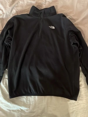 Svart fleece från The North Face - Mjuk och skön svart fleece från The North Face med halv dragkedja och hög krage. Diskret logga på bröstet. Perfekt för lager-på-lager och riktigt chill stil. Materialet är fleece som håller dig varm under kyliga dagar.