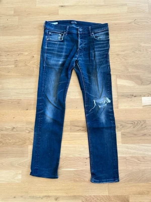 Svarta slitna jeans - Säljer ett par svarta jeans med skinny fit och slitningar på benen, inklusive en patchad detalj på höger lår. Jeansen har klassisk femficksdesign och kontrastsömmar. Hål i ena fickan.