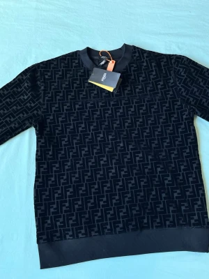 Svart Fendi sweatshirt med logomönster - Snygg svart sweatshirt från Fendi med helöverdraget FF-logomönster. Tröjan har rund halsringning, ribbade muddar och nederkant. Perfekt för dig som gillar exklusiva streetwear-plagg och vill sticka ut med en ikonisk design.