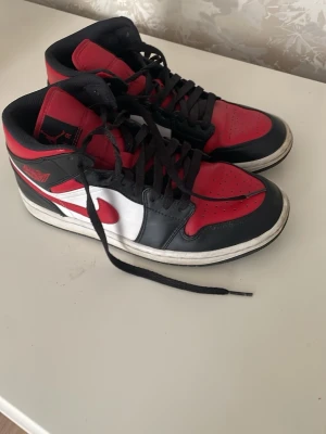 Air Jordan 1 Mid svart/röd/vit - Snygga Air Jordan 1 Mid sneakers i svart, rött och vitt läder. Klassisk design med röd swoosh och Air Jordan-logga på sidan. Skorna har svart snörning och vit sula. Perfekta för dig som gillar streetwear och ikoniska sneakers. De är använda.
