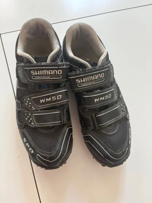 Shimano WM50 cykelskor med kardborre - Storlek 38. Svarta Shimano WM50 cykelskor med vita detaljer och tre kardborreband för smidig justering. Skorna har robust sula med SPD-fäste och är tillverkade i en kombination av syntetmaterial och mesh för bra ventilation. Perfekta för mountainbike eller spinning.