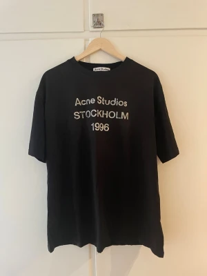 Svart Acne Studios t-shirt med tryck - Svart t-shirt från Acne Studios med vitt tryck på bröstet där det står 'Acne Studios STOCKHOLM 1996'. Klassisk passform med rund hals och korta ärmar. Perfekt för en stilren och trendig look.